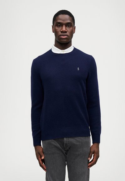 Σκούρο μπλε πλεκτό πουλόβε ρ με στρογγυλή λαιμόκοψη, ριμπ μανίκια και τελείωμα, που φέρει μικρό κεντημένο λογότυπο στο στήθος, φορεμένο πάνω από λευκό πουκάμισο με γιακά.