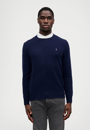 Pull en maille bleu marine avec un col rond, des manches et un ourlet côtelés, arborant un petit logo brodé sur la poitrine, porté par-dessus une chemise blanche à col.