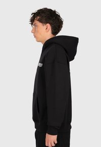 Junger Mann mit lockigem Haar, der einen schwarzen Hoodie mit weißem Text auf der Brust trägt und im Profil vor einem einfarbigen hellen Hintergrund steht.