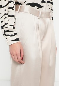 Bézs magasderekú nadrág redőkkel, sima szatén anyagból, hozzáillő övvel. Látható fekete-fehér csíkos hosszú ujjú blúz.