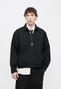 Μαύρο μπουφάν με φερμουάρ και γιακά, με διακριτικό λογότυπο στο στήθος και ελαστικές μανσέτες. Φορεμένο πάνω από λευκό πουκάμισο και γραβάτα.