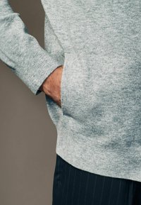Lichtgrijze trui met een gestructureerde afwerking, met een kenguruzak en ribbelmanchetten, gedragen over donker gestreepte broek.