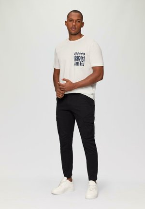 Witte katoenen t-shirt met een "PARADISE" grafiek in marineblauw, gecombineerd met zwarte cargobroeken met zijzakken en witte sneakers.