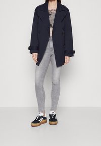 Kobieta w granatowym podwójnym płaszczu, jasnoszarych rurkach, czarno-białych trampkach i wzorzystej bluzce pod płaszczem.