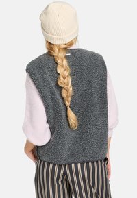 Grijze fleece vest met een gestructureerde afwerking, gedragen over een lichtroze lange mouw top. Een beige gebreide beanie en gestreepte broek maken de look compleet.