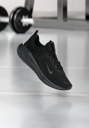 Baskets de sport noires avec une semelle épaisse et le logo Nike swoosh, montrées flottant au-dessus du sol d’une salle de sport avec une barre de poids en arrière-plan.