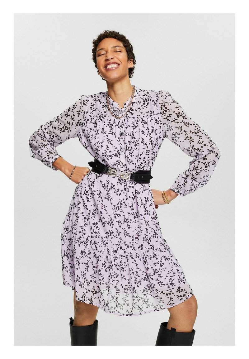 Vestido lila con estampado floral negro, mangas largas, dobladillo con volantes y cinturón negro ajustado. Modelo combinado con botas negras hasta la rodilla.