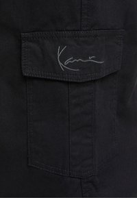 Svart cargoficka med lockdetalj, med en broderad logotyp i grått. Tyget verkar hållbart med en slät textur.