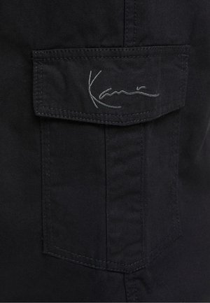 Zwarte cargozak met klepdetail, voorzien van een gestikt logo in grijs. De stof lijkt duurzaam met een gladde textuur.