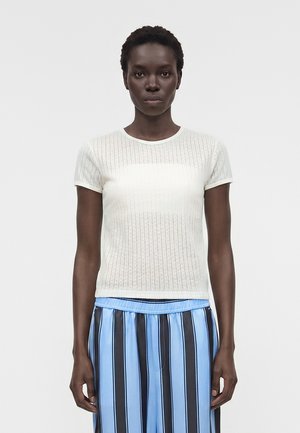 Γυναίκα με κοντά σγουρά μαλλιά φοράει λευκό πλεκτό τοπ με κοντά μανίκια και μπλε παντελόνια φαρδιά με κάθετες μαύρες ρίγες, στέκεται μπροστά σε απλό φόντο.