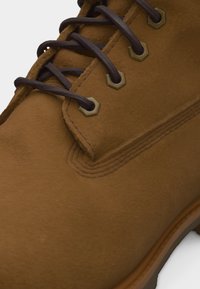 Κοντινή λήψη καφέ μπότες από σουέτ που δείχνει σκούρα καφέ κορδόνια περασμένα μέσα από μπρούτζινες τρύπες και λεπτομερή ραφές κοντά στην μύτη.