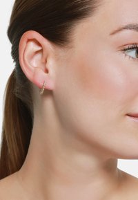 Goldene Creolenohrringe mit kleinen klaren Akzenten, runde Form, polierte Oberfläche, getragen am Ohr eines Models. Haare zurückgebunden, neutraler Hintergrund.