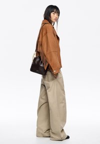 Brązowa zamszowa kurtka z frędzlami, jasnobeżowe oversize'owe spodnie oraz mała czarna torebka z złotym łańcuszkiem.