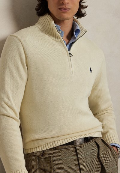 Mees, kes kannab kreemika värvi poolläbi lukuga kudumit sinise kraega särgi kohal, pruuni ruudulise pükstega ja musta vööga.