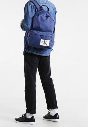 Sac à dos en tissu navy avec une forme ronde, poche frontale zippée et un patch logo blanc. Bretelles visibles et poignée supérieure.