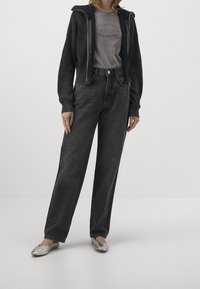 Grau gestrickte Zip-Hoodie mit gerippter Textur, kombiniert mit hochgeschnittenen schwarzen Denim-Jeans. Das Model trägt silberne Ballerinas und hat die Hände lässig platziert.