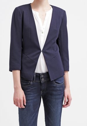 Vrouw draagt een marineblauw blazer met driekwart mouwen over een witte blouse met knopen en donkerblauwe spijkerbroek tegen een egale achtergrond.