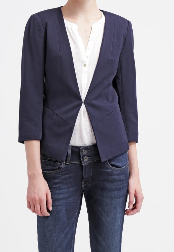 Femme portant un blazer bleu marine à manches trois-quarts par-dessus un chemisier blanc boutonné et un jean bleu foncé sur un fond uni.