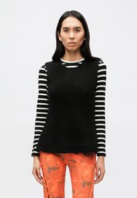 Juodas be rankovių megztinis virš juodo ir balto dryžuoto ilgomis rankovėmis marškinio, derinamas su oranžinėmis raštuotomis kelnėmis. Paprasta fonas.