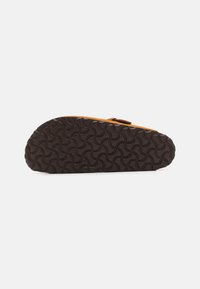 Brun gummisåle med bølgemønster, med en overdel i tan skinn som har en stroppdetalj. Legg merke til den teksturerte og fleksible såleutformingen.