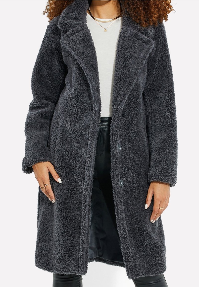 Femme portant un long manteau teddy gris foncé duveteux sur un haut blanc et un pantalon noir, avec des colliers dorés superposés visibles.