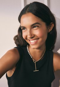 Zlatý náhrdelník s pruhovým príveskom a radom veľkých, lesklých perál. Modelka má na sebe čierny top bez rukávov. Hladká textúra pleti.