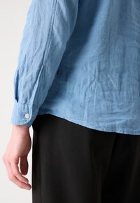 Lichtblauw, gestructureerd overhemd met lange mouwen en met knoopsluitingen, met een iets gekreukt uiterlijk en een gebogen zoom. Zwarte broek zichtbaar onderaan.