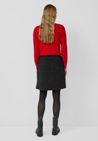 Frau mit langen, welligen Haaren, die einen roten Pullover, einen schwarz gepunkteten Rock, schwarze Strumpfhosen und schwarze Ankle Boots trägt, steht mit dem Rücken zu uns.