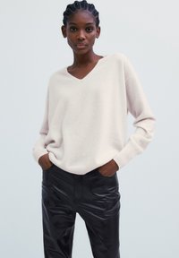 Eine Frau mit geflochtenem Haar trägt einen lockeren, hellbeigen V-Ausschnitt-Pullover und schwarze Lederhosen und steht mit den Händen in den Taschen vor einem schlichten Hintergrund.