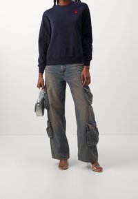 Navy sweatshirt with crew neck and red heart detail, paired with light-wash cargo pants featuring large pockets and a relaxed fit. Silver handbag.
