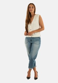 Donna dai lunghi capelli che indossa un gilet bianco senza maniche, jeans blu arrotolati e tacchi alti neri, in piedi davanti a uno sfondo bianco.