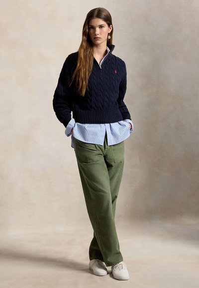 Tânără cu păr lung și șaten, purtând un pulover bleumarin cu model tricotat peste o cămașă albastră cu dungi, pantaloni verzi, adidași albi, stând cu mâinile în buzunare.