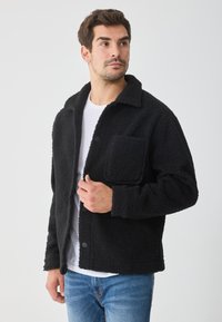 Juodas fliso švarkas su tekstūruotu paviršiumi, apykakle ir priekinėmis kišenėmis. Jis turi užsegimus mygtukais ir yra dėvimas ant baltos marškinėlių.