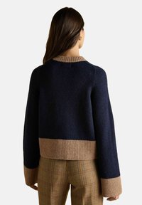 Žena v tmavomodrom svetri s béžovými manžetami a lemom, v kombinácii s hnedými kockovanými nohavicami, zobrazená zozadu.