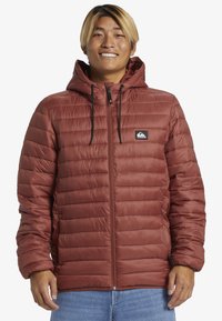 Rostfarvet pufferjakke med hætte, som har lynlås foran, kviltet design, justerbare snore og et logo patch på brystet.