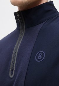 Uomo che indossa una giacca blu navy con zip e collo alto, con il logo "B" in rilievo sul petto, zip parzialmente chiusa vicino al collo.