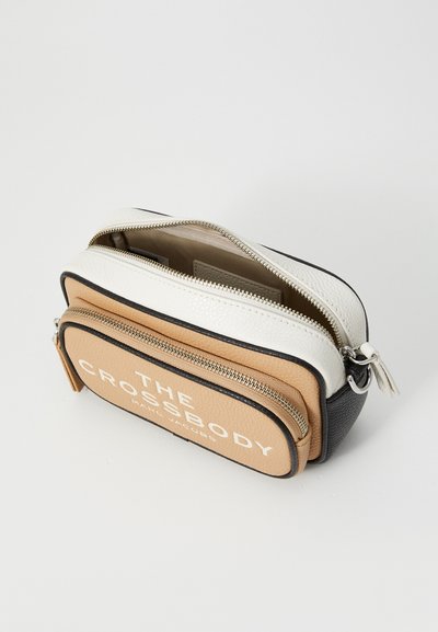 Kompakt tre-farget skinn crossbody-veske med doble glidelåslommer, beige frontlomme merket "THE CROSSBODY," og sorte, hvite, og brune fargeblokker.