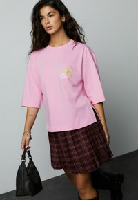 Lyserød oversized t-shirt med korte ærmer, der har broderet "Slow Down" tekst og blomsterdesign. Kombineret med et ternet plisseret skørt og håndtaske.