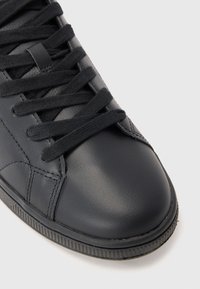 Sneaker de pele preta com atacadores pretos, costura visível e sola de borracha texturizada contra um fundo branco.