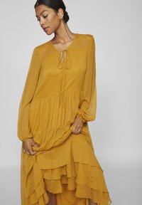 Sundress longa de cor mostarda, feita de um tecido leve, com um fundo franzido, mangas longas e um detalhe de amarração no pescoço, apresentando uma silhueta fluida.