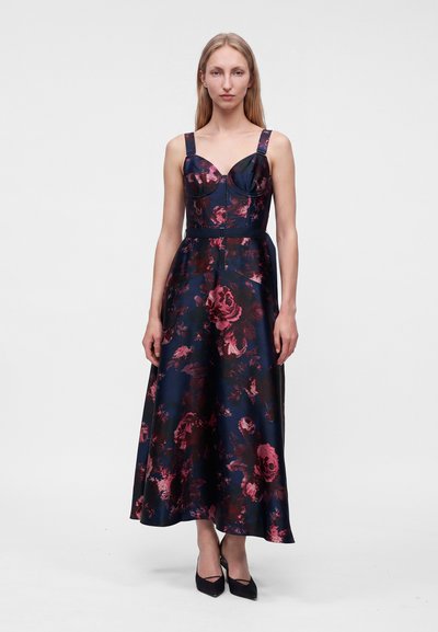 Femme portant une robe midi bleu foncé sans manches avec un imprimé floral rose et des escarpins noirs à bout pointu, debout devant un fond uni.