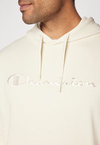 Krémová mikina s kapucňou vyrobená z mäkkého materiálu má výrazné vyšívané logo "Champion" vo svetlo béžovej farbe a nastaviteľné šnúrky.