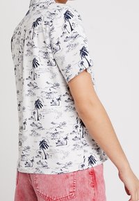 Košile na knoflíky s krátkým rukávem ve světle šedé látce s navy potiskem palem a krajiny, s klasickým límcem a volným střihem.