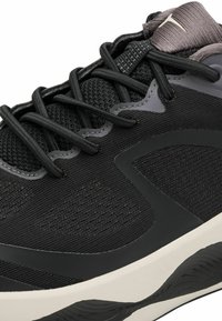 Black athletic shoe features a textured upper with mesh and synthetic overlays, contrasting rubber midsole, and black lace detailing.