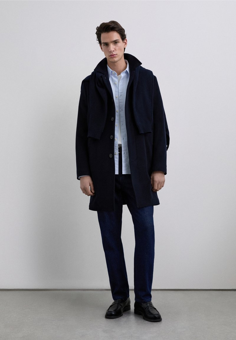 Tmavomodrý vlněný kabát s vysokým límcem, tmavě modré džíny a černé kožené boty. Košile je světle modrá s knoflíky. Obsahuje svetr přehozený přes kabát.