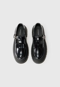 Chaussures en cuir noir avec une semelle épaisse, présentant un devant plat, des accents de sangle et un détail de boucle en argent. Texture lisse avec brillance.