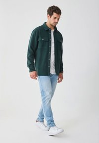 Camicia di flanella verde scuro con bottoni e due tasche sul petto, indossata sopra una t-shirt bianca, abbinata a jeans azzurri e scarpe da ginnastica bianche.
