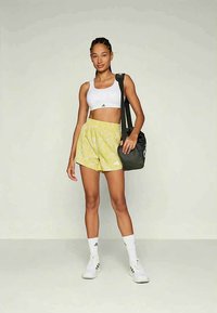 Αθλητική γυναίκα με λευκό σπορ σουτιέν, κίτρινες βερμούδες με σχέδιο, λευκές κάλτσες και αθλητικά παπούτσια, κρατώντας μαύρη τσάντα γυμναστηρίου στον έναν ώμο.