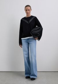 Femme debout portant un pull noir avec des détails en dentelle, un jean large bleu clair, des chaussures noires et tenant un grand sac en cuir noir.