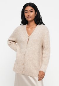 Cardigan fluffos, roz deschis, cu decolteu în V, mâneci lungi și cinci nasturi pe față. Textură moale cu margine și mansete cu filet.